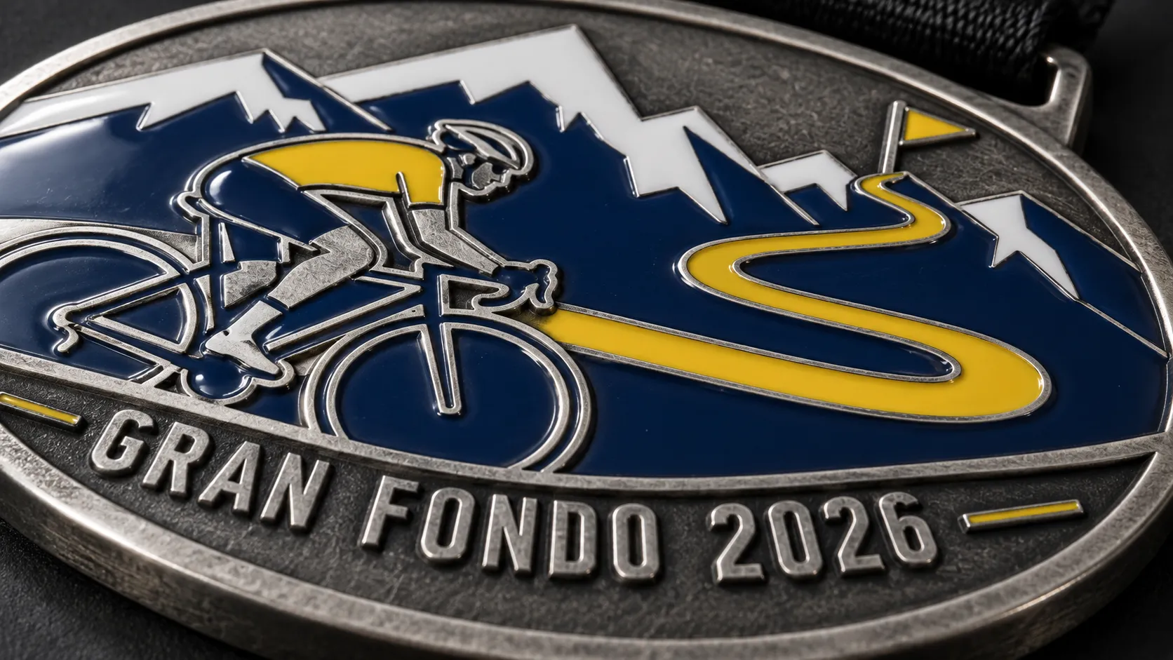 Macro of a cast cycling medal: a hairpin road in yellow enamel climbing towards navy mountains, with GRAN FONDO 2026 lettering