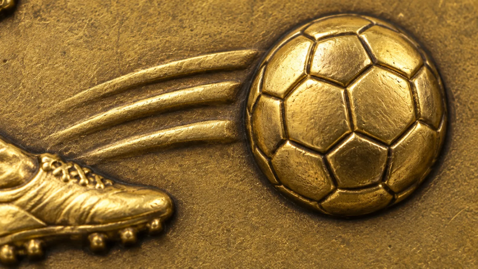 Macro of a cast football medal: the ball and a football boot in antique gold relief, every panel and stud visible